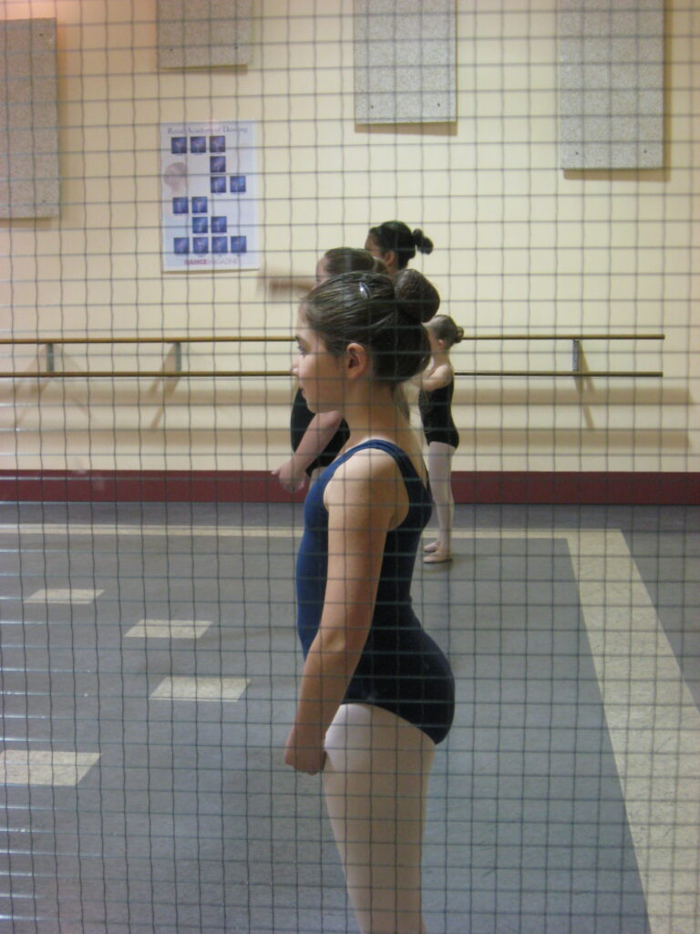 Author as a preteen in a dance workshop. She is standing profile to the camera wearing a navy blue leotard and pink tights. Her dark hair is in a high bun. She stands with her arms in bras bas, and feet in 1st position.